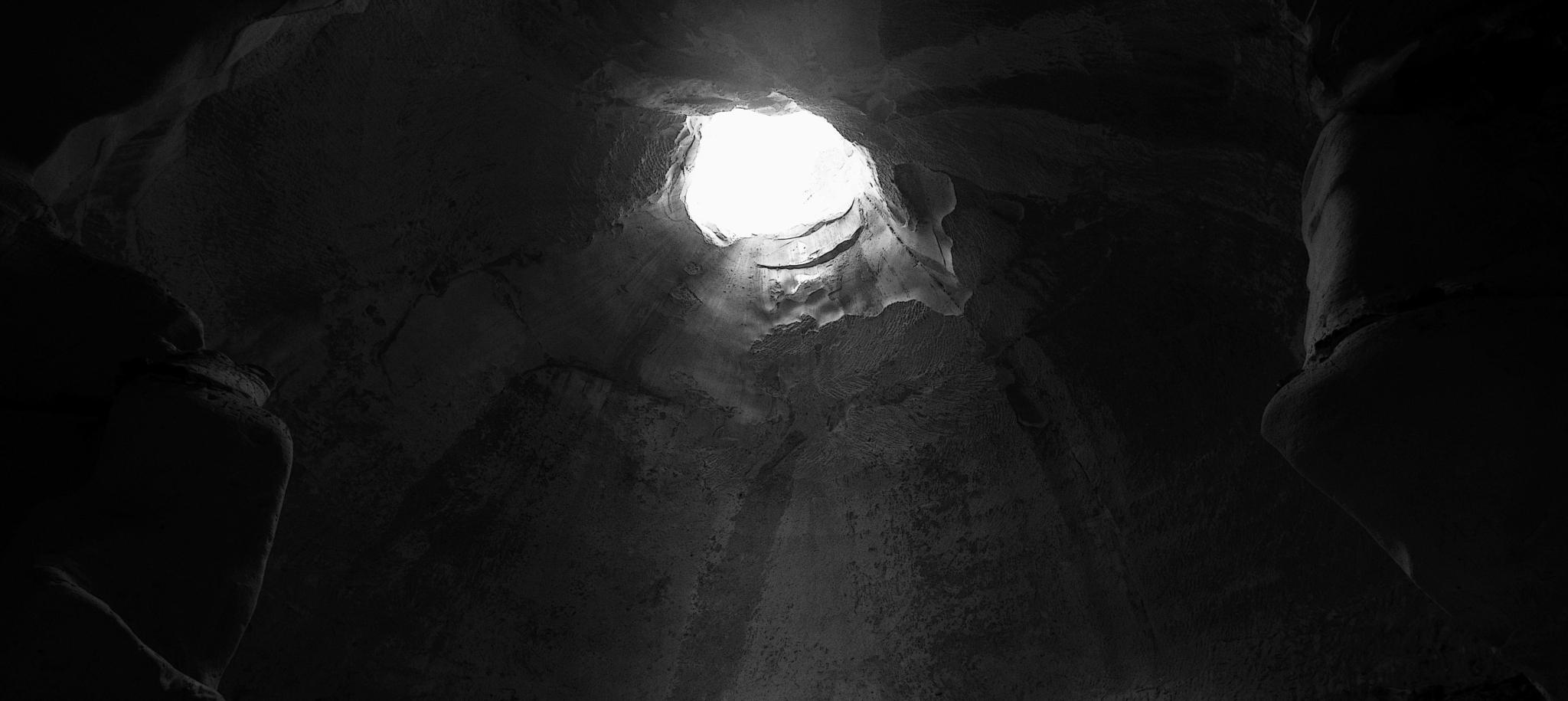 A lone figure stands in a cavernous space, illuminated by a circular opening above, creating a dramatic interplay of light and shadow.